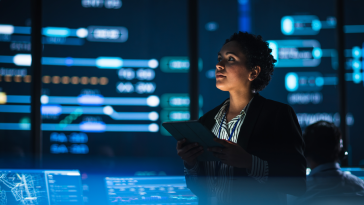 Young Female Government Employee Uses Tablet Computer in System Control Monitoring Center. In the Background Her Coworkers at Their Workspaces with Many Displays Showing Technical Data.
